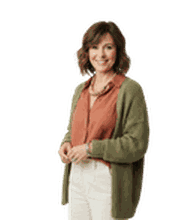 Smiling woman with curly hair, wearing a rust-colored shirt, white pants, and green cardigan, standing against a plain white background.