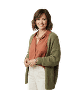 Smiling woman with curly hair, wearing a rust-colored shirt, white pants, and green cardigan, standing against a plain white background.