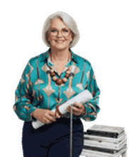 Smiling woman with glasses wearing a teal blouse holds rolled papers and stands beside stacked magazines.