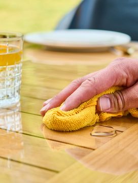Hand wiping a wooden table with a yellow cloth, near a glass of orange beverage and a ring, in an outdoor setting.