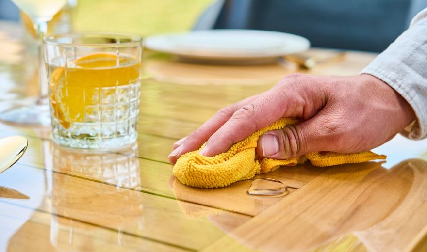 Hand wiping a wooden table with a yellow cloth, near a glass of orange beverage and a ring, in an outdoor setting.