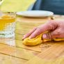 Hand wiping a wooden table with a yellow cloth, near a glass of orange beverage and a ring, in an outdoor setting.