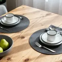 A wooden table set for two with white dishes, cups, and cutlery on gray placemats. A bowl of green pears is nearby.