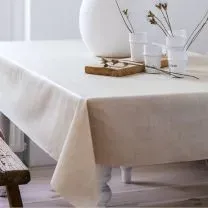 A table with a beige tablecloth, a white vase, three glasses, and a wooden tray with twigs, set in a bright room.