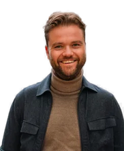 Smiling man with a beard, wearing a brown turtleneck and dark jacket, standing against a blurred background.