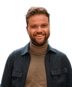 Smiling man with a beard, wearing a brown turtleneck and dark jacket, standing against a blurred background.