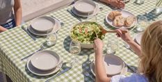 A table set with white plates, glasses of water, sliced bread, and a large bowl of salad on a green checkered tablecloth. People are seated nearby.