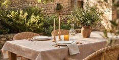 Outdoor dining setup with a linen-covered table, wicker chairs, plates, candles, a glass carafe, and a plant in a terracotta pot against a stone wall background.