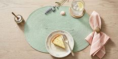 Een stuk cake op een bord met een vork op een groen placematje. Een glas citroenwater, een gestreept servet, lavendel en een suikerpotje ernaast.