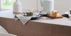 A table with a brown tablecloth set for breakfast, featuring a mug, a pastry on a wooden board, a teapot, and a white vase.
