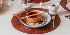 A table setting with a brown placemat, stacked plates, an orange bowl, a breadstick, a gold spoon, and a cup of frothy coffee.