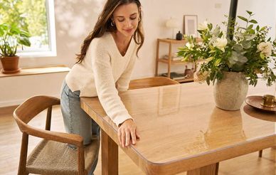 A woman in a cozy sweater leans on a wooden table, lightly touching its surface, with a vase of white flowers and greenery next to her.