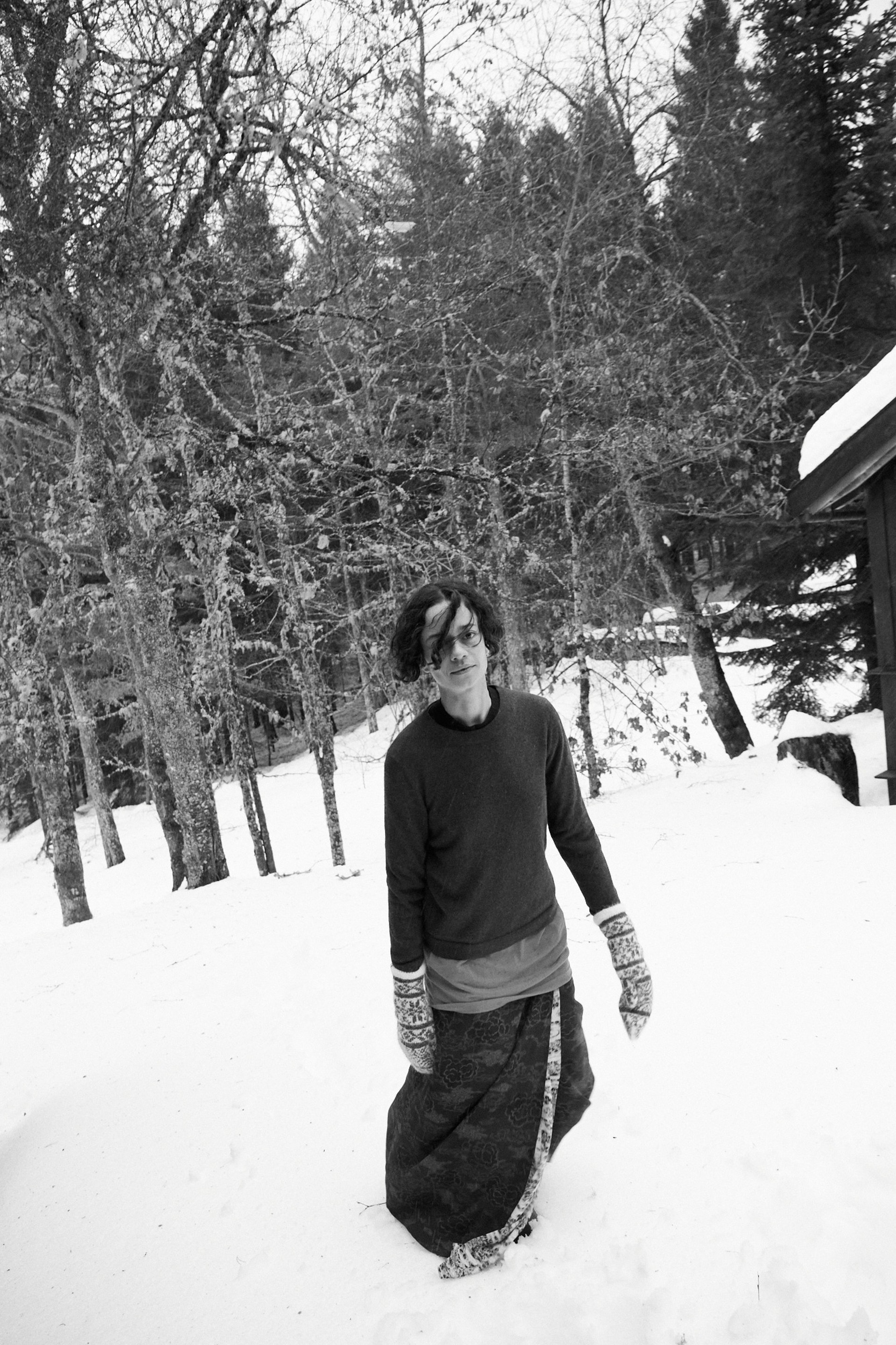 Person with long hair stands outdoors, looking to the side, with a blurred landscape in the background.