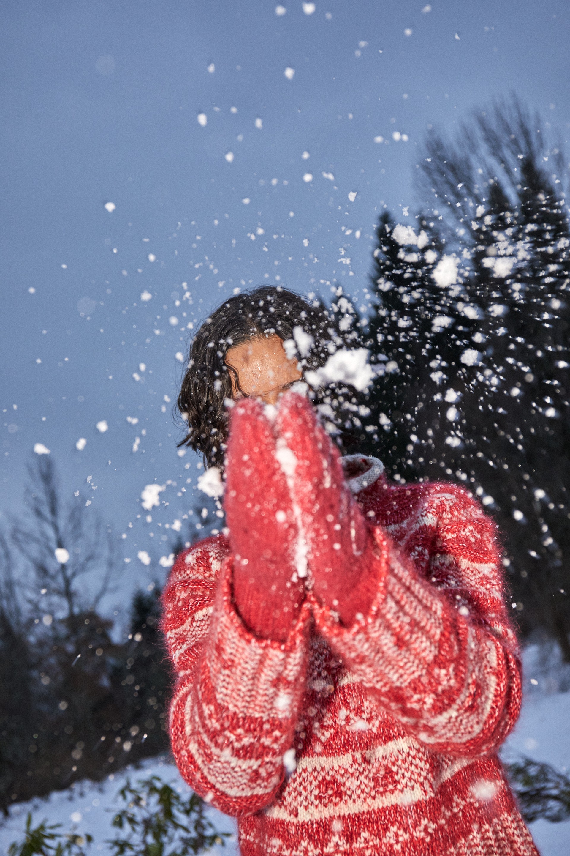 Woman in a knitted vest and casual attire sits on a boat, with water and greenery in the background.