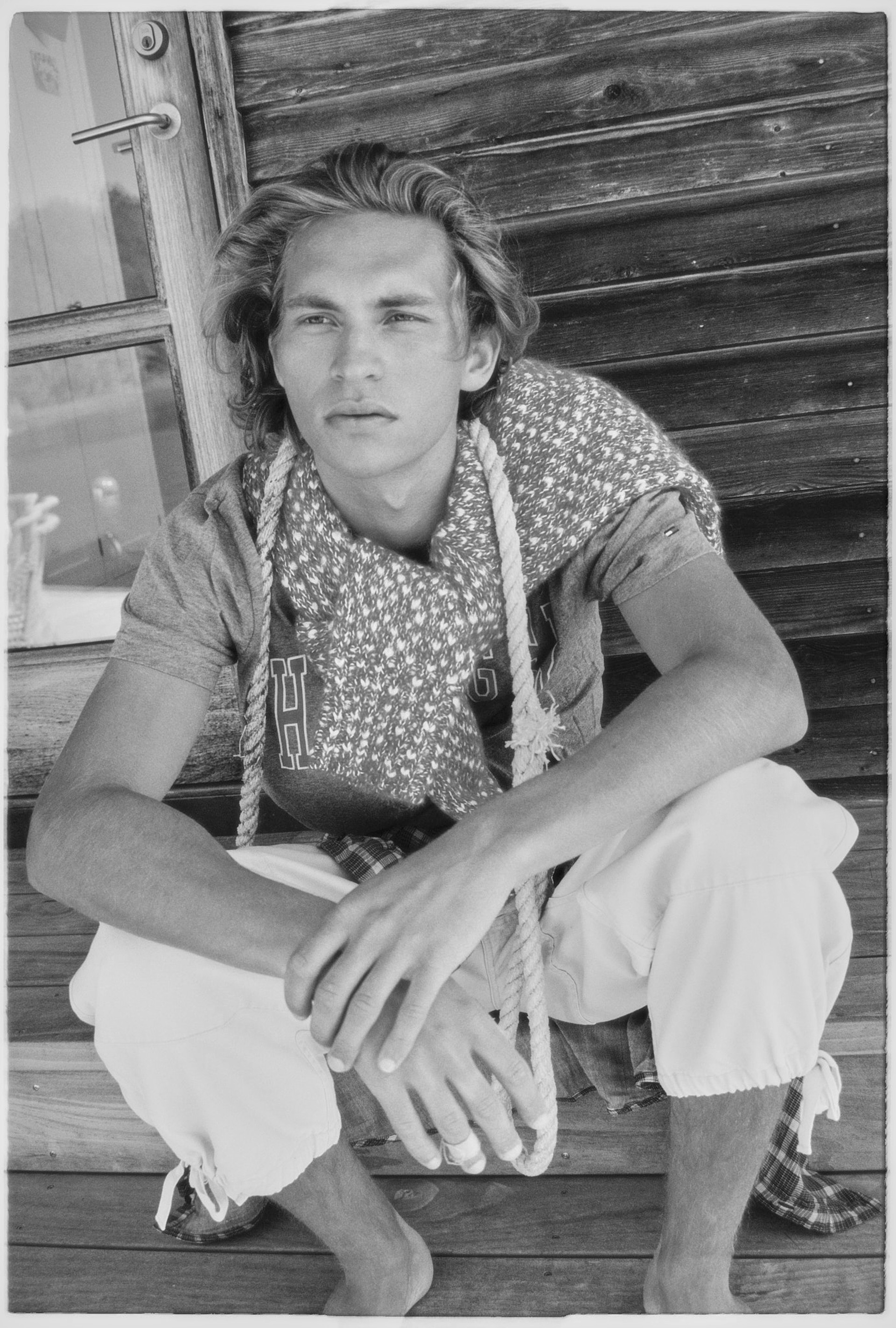 Person in a beanie and sweater sits on a rock by a rocky shoreline, looking at the camera under a clear sky.