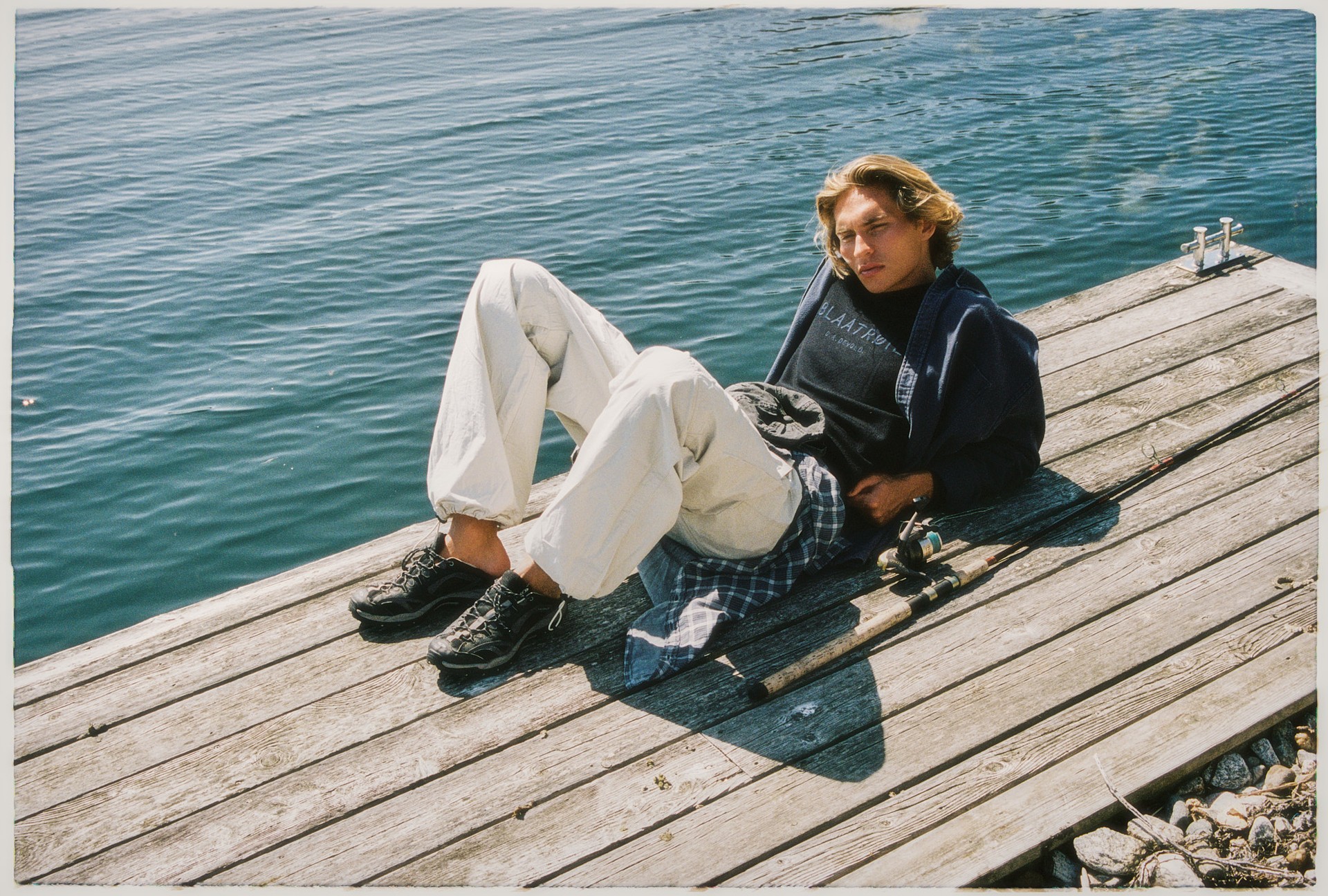 Person standing in a grassy landscape with hills in the background, wearing a dark coat and white hoodie. Wind is blowing their hair.