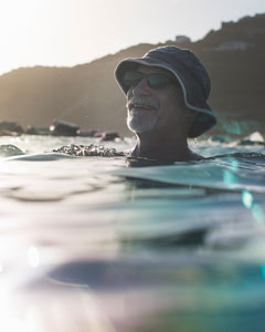 Person mit Sonnenhut und Sonnenbrille befindet sich im Wasser nahe einer Küste. Im Hintergrund sind Hügel und das Ufer zu sehen.