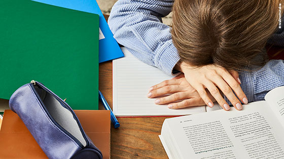 Person sitzt zwischen Schul- und Lernunterlagen mit dem Kopf auf den Armen am Schreibtisch.