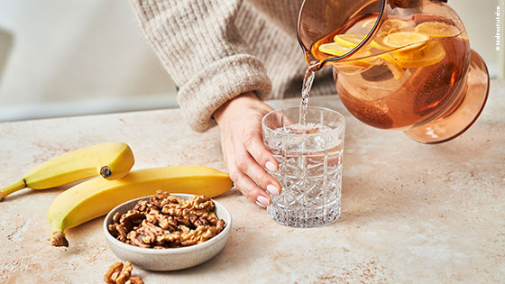Zitronenwasser wird ein ein Glas gefüllt, nebenbei zwei Bananen und eine Schale mit Nüssen.