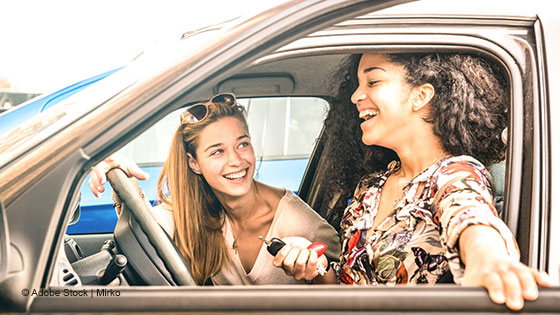 Zwei Personen sitzen in einem Auto, während eine Person der anderen einen Autoschlüssel übergibt.