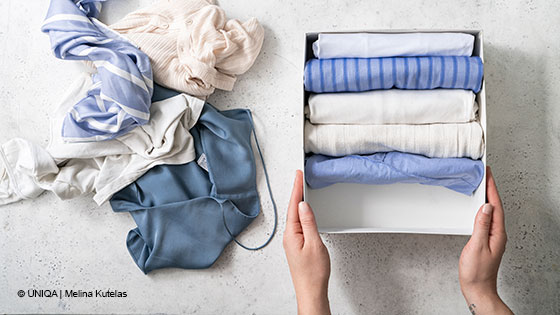 Hände halten eine Box mit ordentlich gefalteter Kleidung. Links daneben liegt ein Stapel ungefalteter Kleidungsstücke