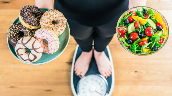 Person auf der Waage. In den Händen hält sie einen Teller mit Donuts und einen Teller mit Salat