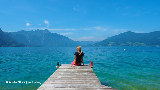 Frau sitzend am Steg und schaut auf den weiten See. Strahlender Sonnenschein, Sommer, im Hintergrund Berge.