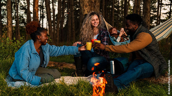 Eine Freundesgruppe mit Tassen am Lagerfeuer, im Hintergrund eine Hängematte und Wald.