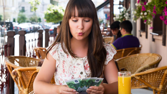 junge Dame sitzt auf Strohsesseln vor einem Lokal, trinkt Orangensaft und zählt Geld