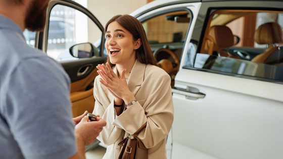 Frau steht vor freudig vor ihrem Auto. Ein Mann reicht ihr den Autoschlüssel.
