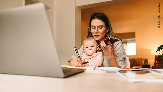 Frau mit Baby sitzt vor dem Laptop und telefoniert.