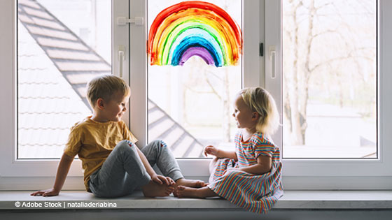 Zwei Kinder sitzen auf einer Fensterbank vor einem Fenster mit einem gemalten Regenbogen.