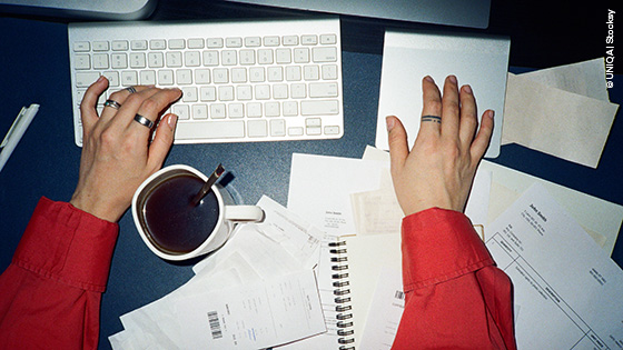 Hände auf einer Tastatur und Touchpad. Nebenbei eine Tasse Kaffee und verschiedene Papiere