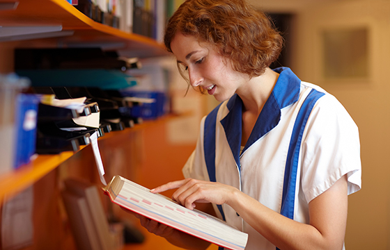 Eine Frau in einer weiß-blauen Uniform liest eine Akte in einem Büro, im Hintergrund befinden sich Regale und Dokumente.