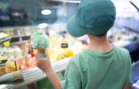 Kind mit einem Eis in der Hand begutachtet eine Eistruhe mit vielen verschiedenen Sorten