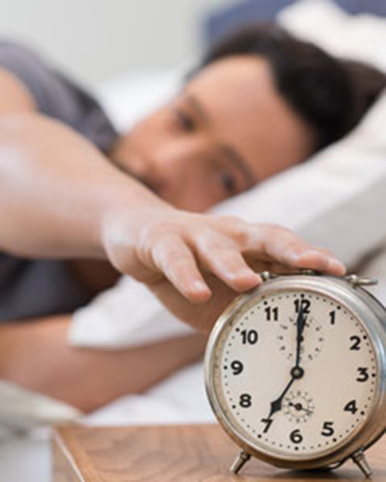 Person liegt im Bett und greift nach einem Wecker auf einem Nachttisch.