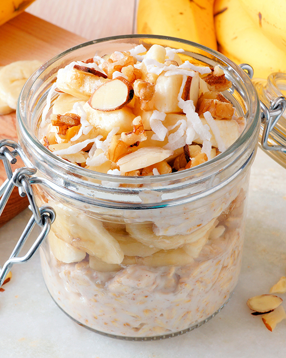Glas mit Haferflocken, Bananenscheiben, Nüssen und Kokosraspeln auf einer hellen Oberfläche.