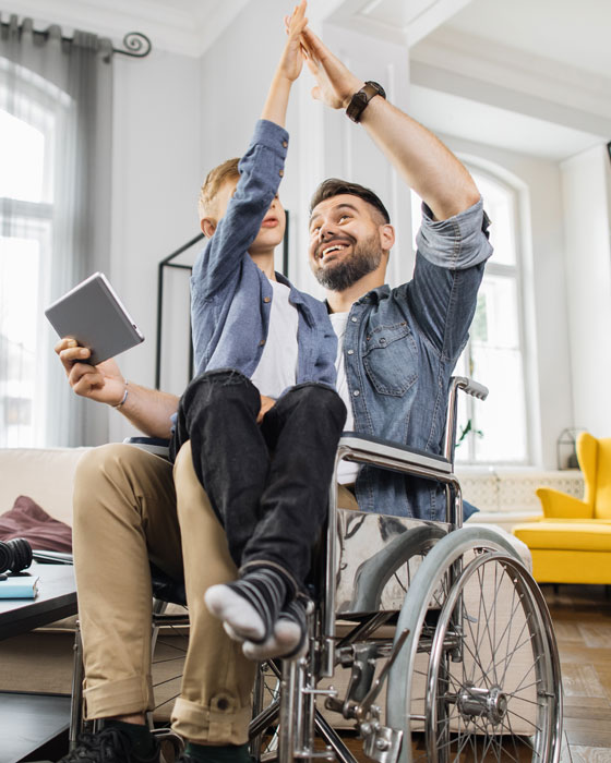 Person sitzt in einem Rollstuhl in einem Wohnraum und hält ein Tablet in der Hand. Ein Kind sitzt auf dem Schoß. Beide heben die Hände nach oben.