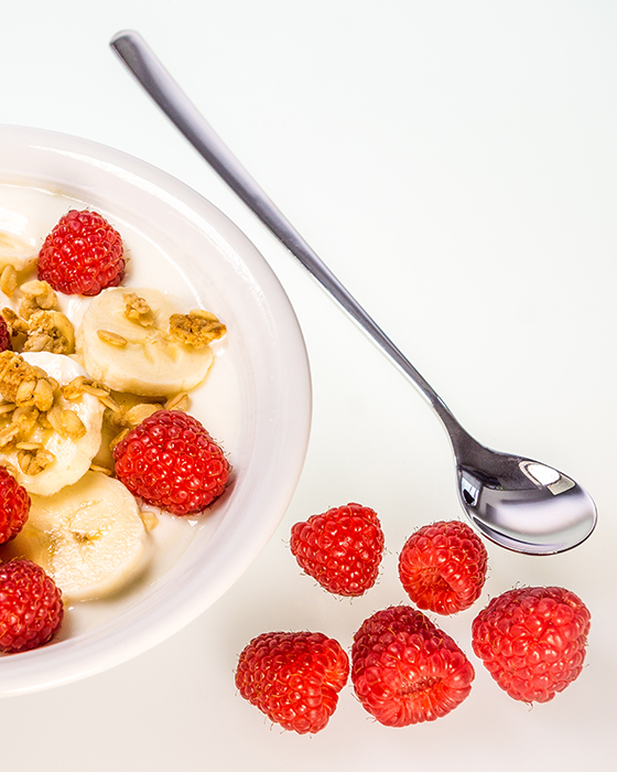 Schale mit Joghurt, Bananenscheiben, Himbeeren und Müslimischung neben einem Löffel.