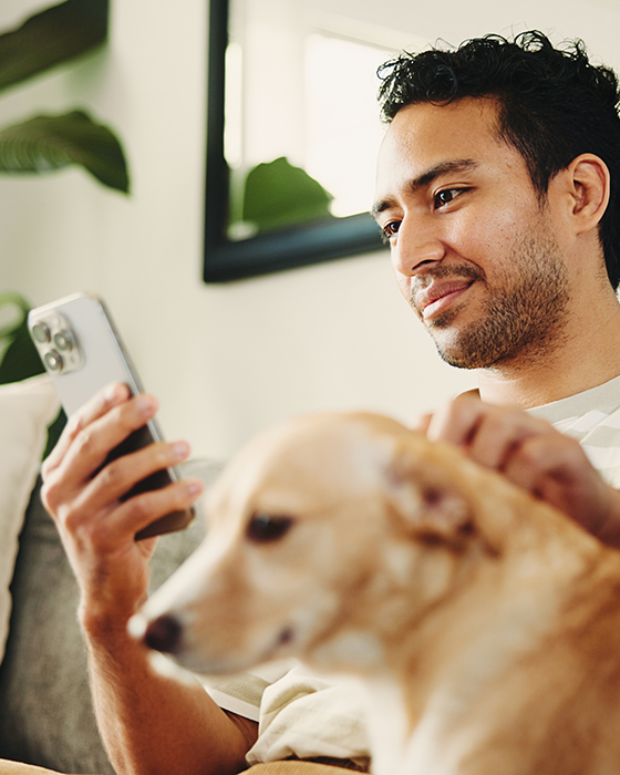 Person sitzt mit einem Hund auf einem Sofa und schaut auf ein Smartphone.