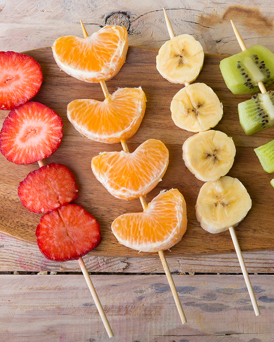 Obststücke aus Erdbeeren, Mandarinen, Bananen und Kiwis auf Holzspießen, angerichtet auf einem Holzbrett