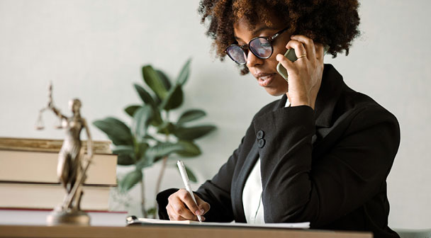 Dame am Telefon notiert etwas auf einen Zettel, daneben steht Justitia in Form einer Miniaturstatue auf dem Tisch