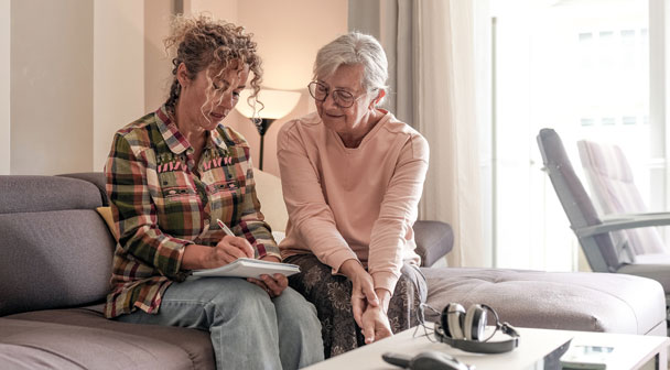 Pflegekraft und Patientin sitzen auf der Couch und notieren Infos auf einen Notizblock