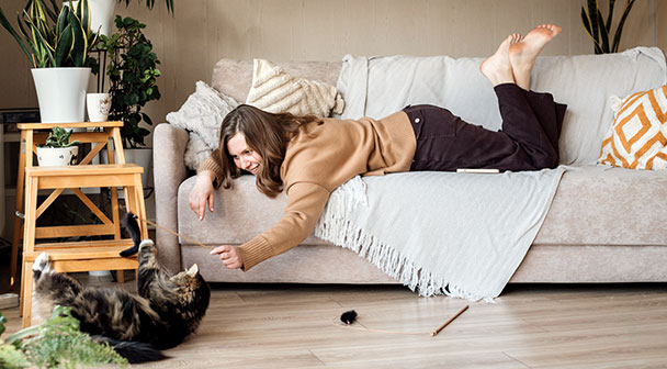 Eine Frau liegt gemütlich am Sofa im Wohnzimmer und spielt mit der Katze