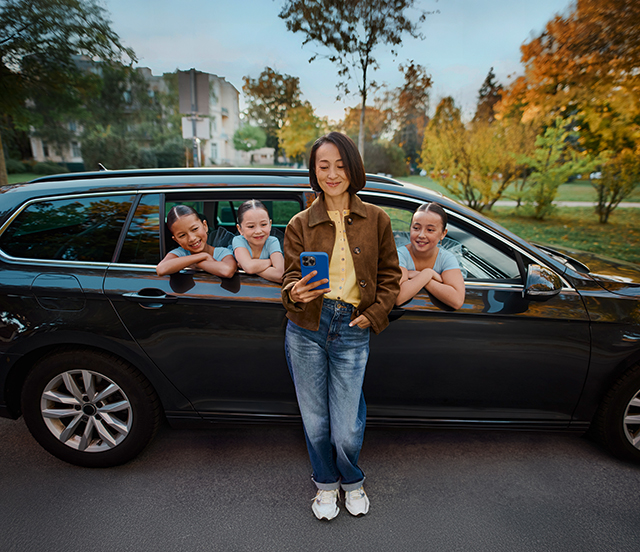 Mama lehnt sich auf das Smartphone schauend an ihr Auto. Die Kinder sitzen im Auto und schauen aufs Handy.