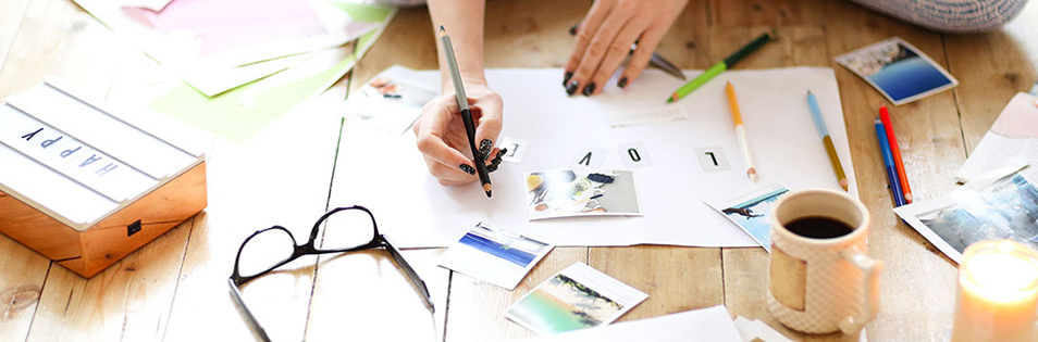 Hände gestalten ein Vision Board auf einem Tisch mit Fotos, Papierkarten, Stiften und einer Tasse.