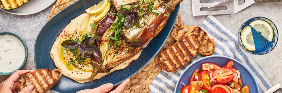 Gegrilltes Gemüse, Brot und ein Tomatensalat sind auf einem gedeckten Tisch angerichtet. Daneben stehen ein Getränk und eine Schale mit Dip.