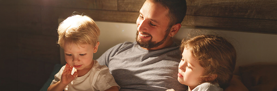 Eine erwachsene Person sitzt mit zwei Kindern im Bett und liest gemeinsam ein Buch in einer gemütlich beleuchteten Umgebung
