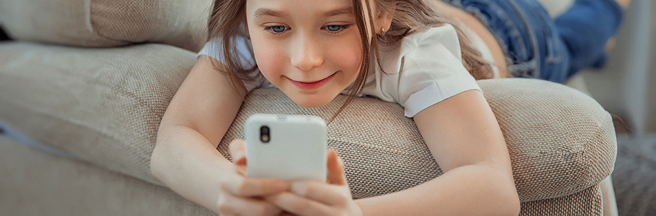 Kind liegt auf einem Sofa und hält ein Smartphone in beiden Händen.