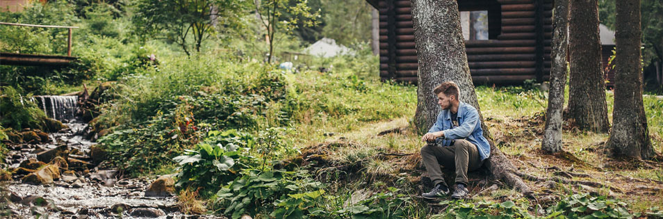 Person sitzt am Ufer eines kleinen Bachs in einer bewaldeten Umgebung. Im Hintergrund steht eine Holzhütte zwischen Bäumen und Pflanzen.